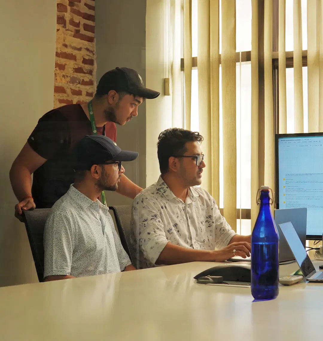 Three individuals collaborating, one standing, two seated, sharing a laptop screen, fully engaged in their shared work.